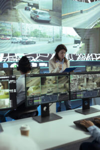 Security operations center with staff monitoring live traffic surveillance footage on multiple screens, while a supervisor reviews notes on a clipboard.