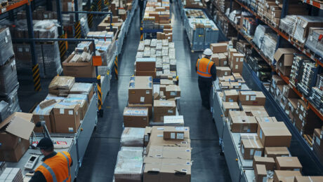 photorealistic-scene-with-warehouse-logistics-operations Warehouse workers in safety vests managing logistics and packaging operations among stacked boxes and inventory shelves in a large distribution center.